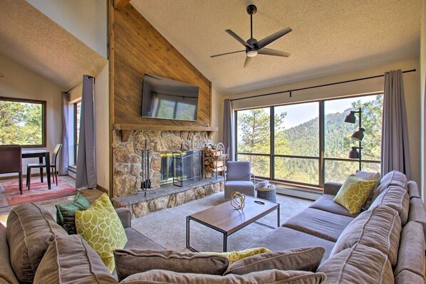 A spacious living room with a stone fireplace, a ceiling fan, and large windows offering a view of the outdoors. - The Evergreen Gem| Mountain Experience Retreat (Evergreen)
