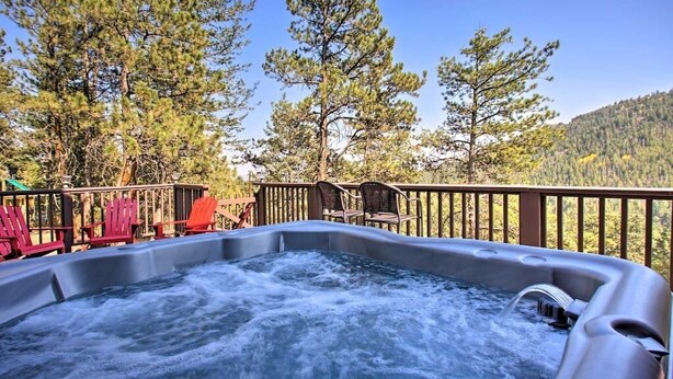A hot tub surrounded by Adirondack chairs and a wooden deck, with a forested mountain backdrop. - The Evergreen Gem| Mountain Experience Retreat (Evergreen)