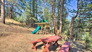 A picnic area with two red picnic tables and a playground with a slide in a forested setting. - The Evergreen Gem| Mountain Experience Retreat (Evergreen)