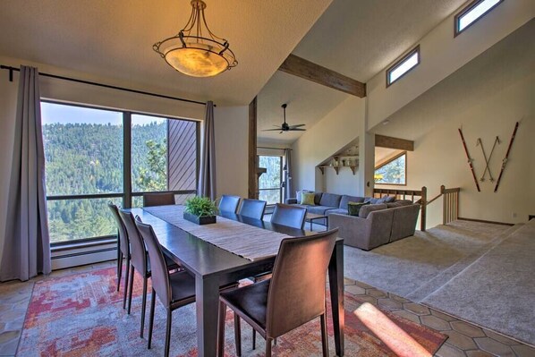 A spacious living area with a dining table, chairs, and a sofa. There is a large window offering a view of a forested area.