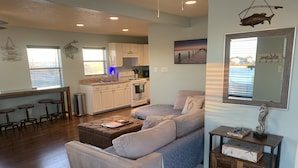 A modern living room with a sofa, ottoman, and a small table with decorative items. A kitchen with white cabinets and a dining area with bar stools is visible in the background. - Sunny San Leon Casita!! (San Leon)