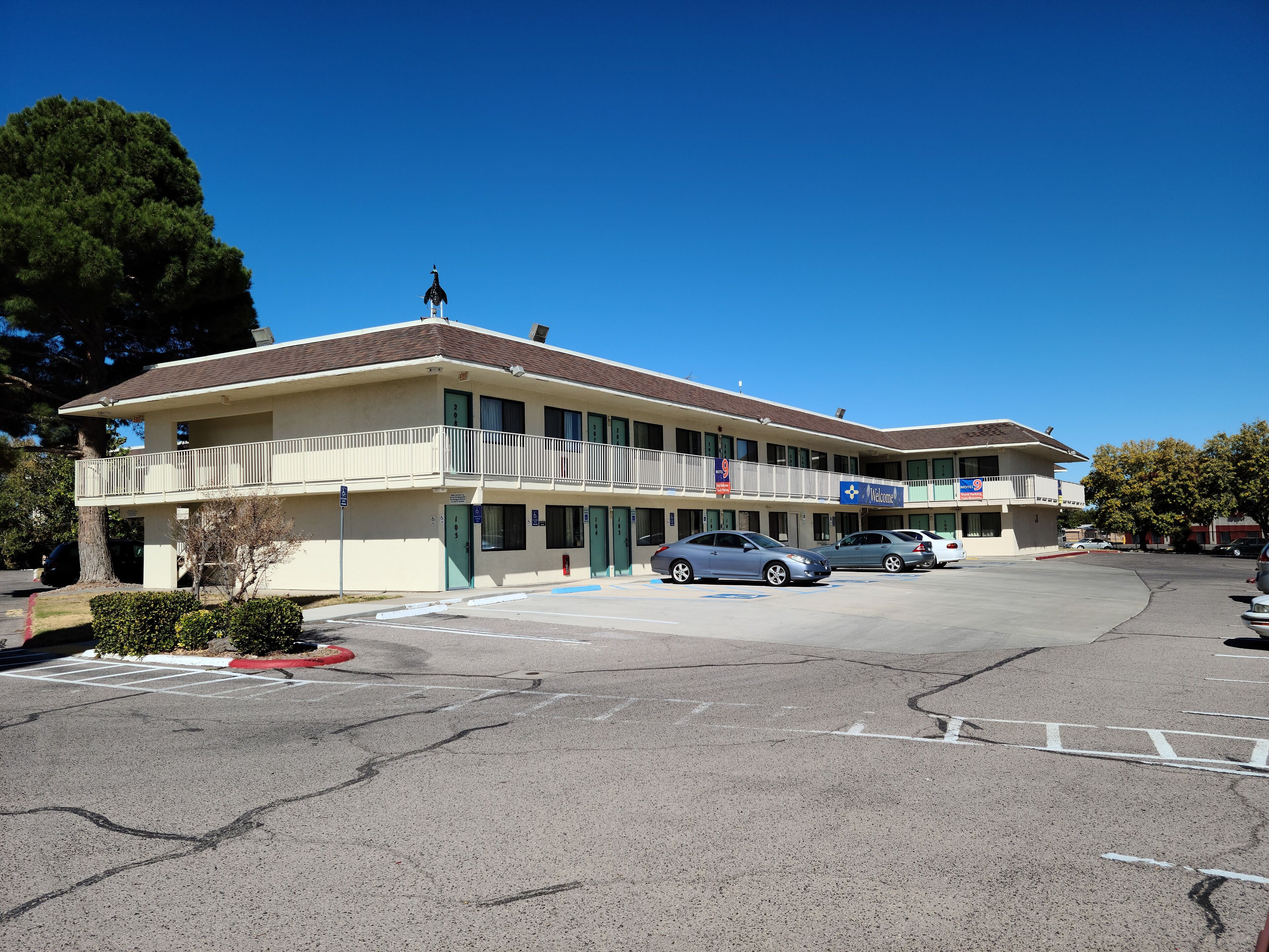 A multi-story motel with a flat roof, a balcony on the second floor, and a parking lot with several cars.