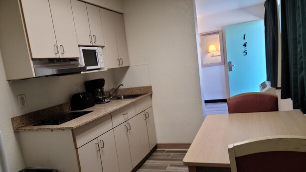 A compact kitchen with a microwave, coffee maker, and a small trash bin. A dining table and chairs are visible in the foreground. - Motel 9 Las Cruces (Las Cruces)