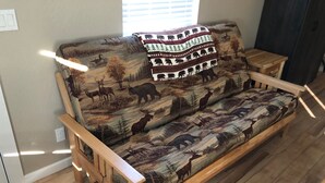 A wooden sofa with a wildlife print, a small wooden side table, and a window with blinds. - Riverside Cabin 2 - (Brand New Construction) (Grants Pass)