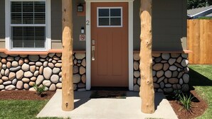 A house with a brown front door, a stone foundation, and a small porch with two wooden pillars. - Riverside Cabin 2 - (Brand New Construction) (Grants Pass)