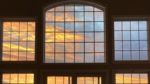 A room with large windows overlooking a body of water, a patterned armchair, a small table with a lamp, and a view of the sunset. - Relax lakeside at our spacious Geneseo house on  Conesus Lake (Conesus)