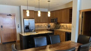 A modern kitchen with wooden cabinets, a stainless steel refrigerator, a microwave, and a granite countertop. There is a dining table with chairs and pendant lighting above.