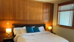 A hotel room with a large bed, wooden headboard, two bedside lamps, and a window with blinds.