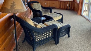 A log cabin interior with two wicker chairs, a lamp, and large windows.