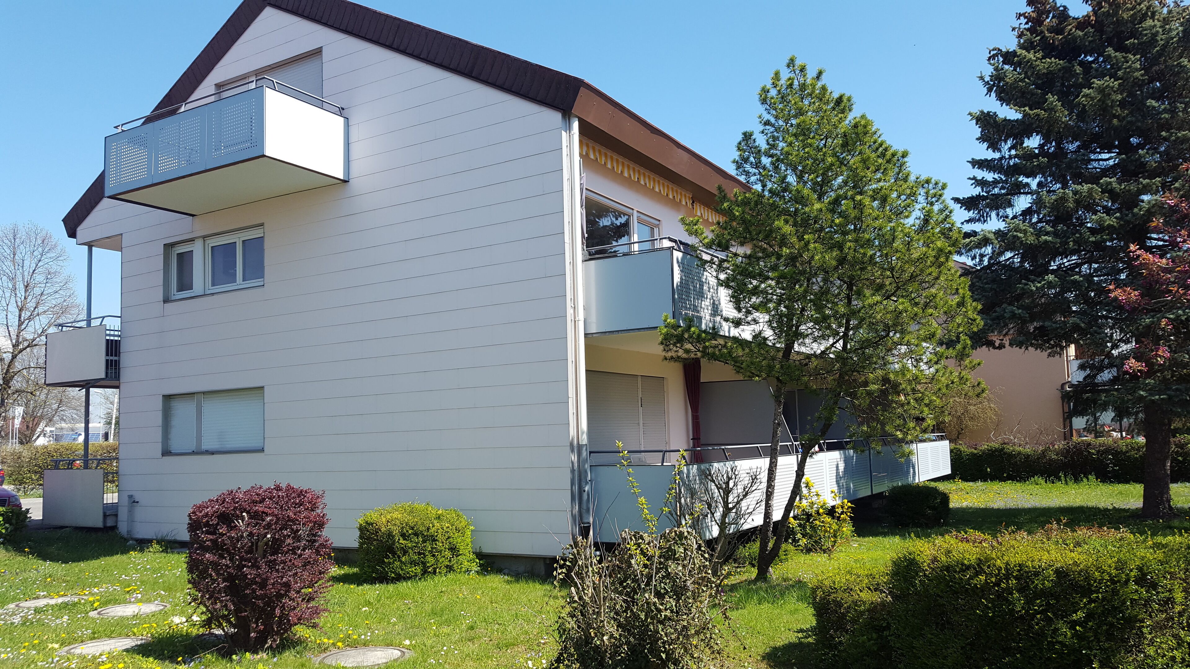 A two-story residential building with a balcony on the upper floor, surrounded by greenery and trees.