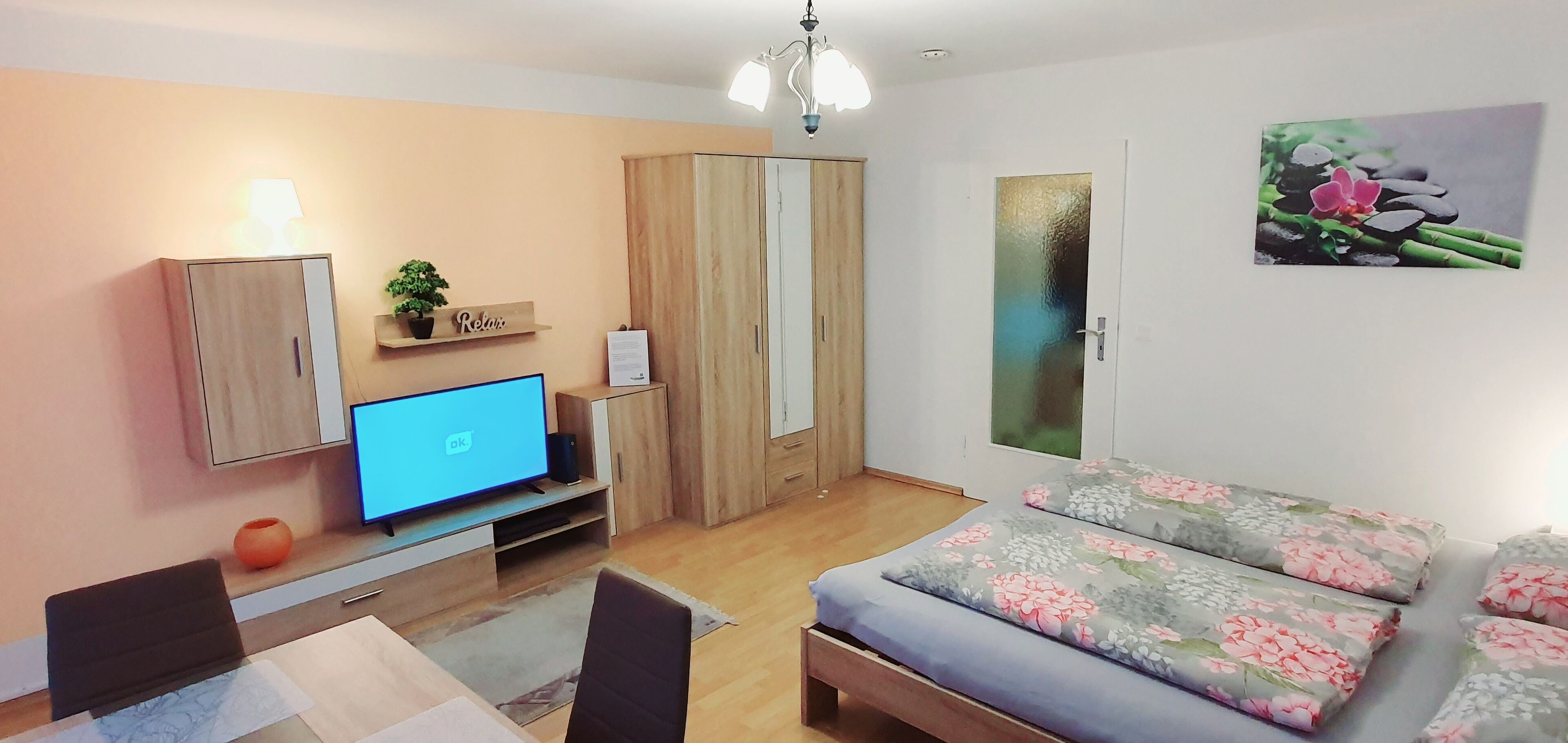 A modern living room with a flat-screen TV, wooden furniture, a sofa with floral cushions, and a wall-mounted artwork.