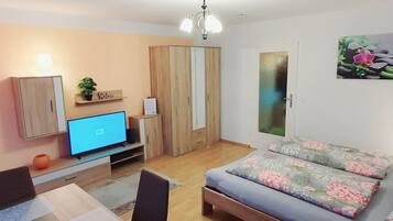 A modern living room with a flat-screen TV, wooden furniture, a sofa with floral cushions, and a wall-mounted artwork.