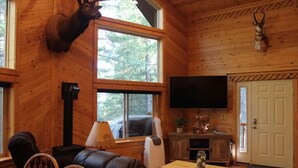 A cozy cabin interior with a fireplace, a mounted deer head, a television, and a dining area. - Mountain Pines Luxury Yellowstone Cabin + Hot Tub + Sleeps 15 + Garage + 2 Acres (Island Park)