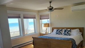 A bedroom with a wooden bed, a ceiling fan, a view of the beach, and a window with a view of the ocean.