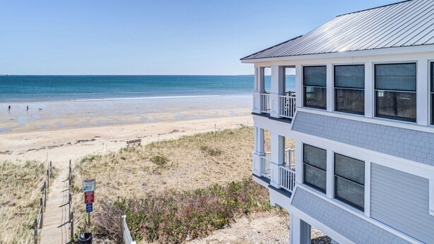 A beachfront property with a balcony overlooking the ocean, a sandy beach, and a clear sky.