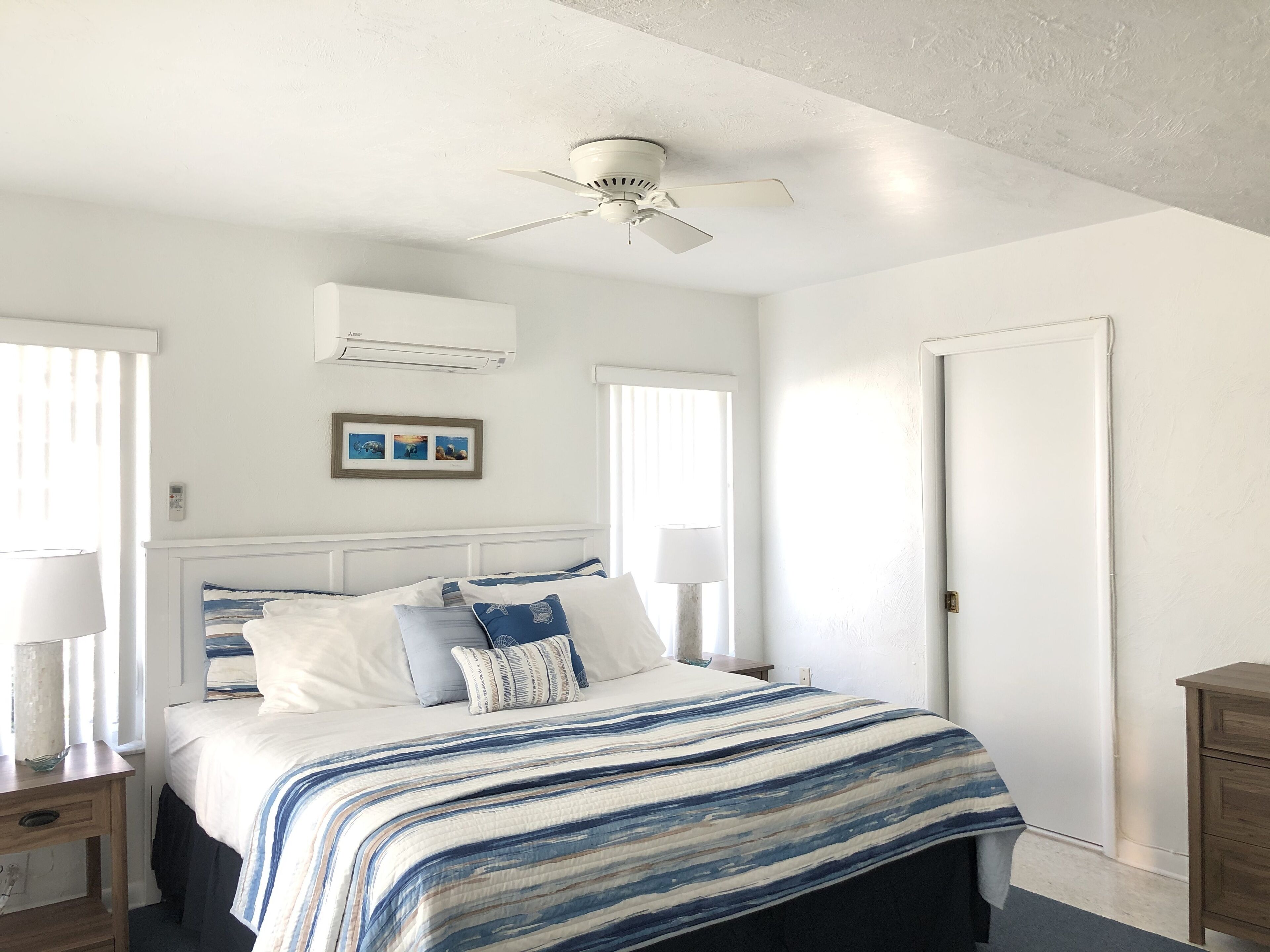 A bedroom with a bed, bedside lamps, a ceiling fan, a framed picture, and a door.