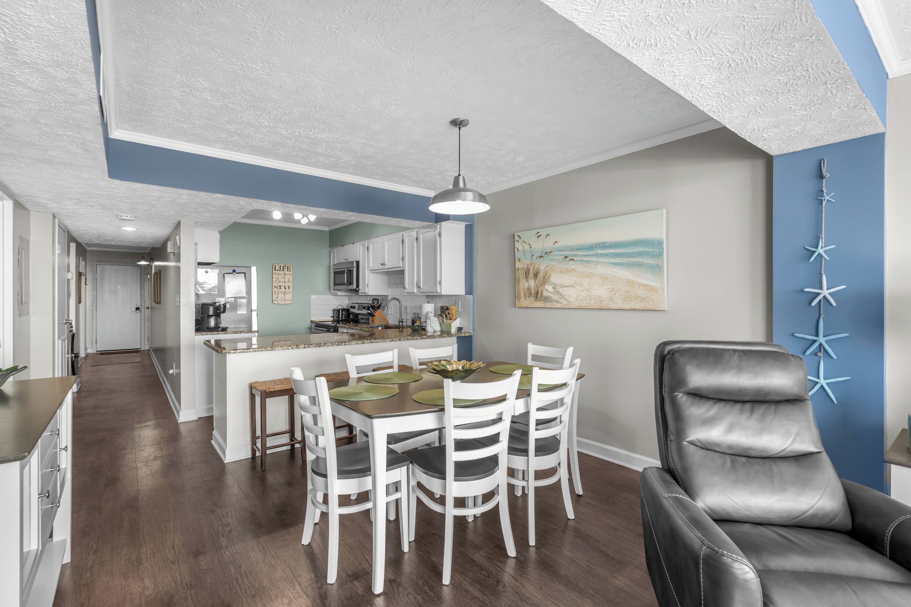 A modern kitchen with white cabinets, a central island, and a dining area with white chairs and a black leather chair.