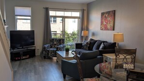 A modern living room with a flat-screen TV, a sofa, a dining table with chairs, and a large window with curtains.