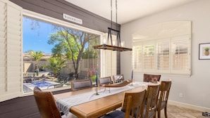 A dining area with a wooden table and chairs, a hanging light fixture, and a view of the backyard through large windows. - Ultimate AZ Getaway! Enjoy this Stunning 5BD with refreshing Pool & Spa! (Goodyear)