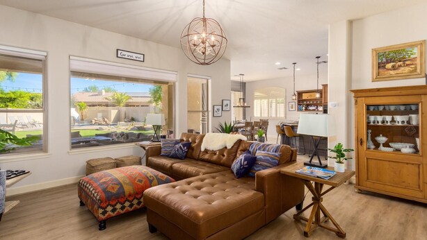 A spacious living room with a brown leather sectional sofa, a wooden coffee table, and a chandelier. There is a dining area with a white table and chairs, and a kitchen visible in the background. - Ultimate AZ Getaway! Enjoy this Stunning 5BD with refreshing Pool & Spa! (Goodyear)