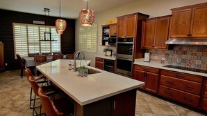 A modern kitchen with a central island, wooden cabinets, stainless steel appliances, and a decorative backsplash. - Ultimate AZ Getaway! Enjoy this Stunning 5BD with refreshing Pool & Spa! (Goodyear)