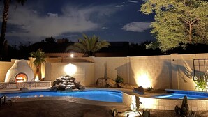 A nighttime outdoor setting with a swimming pool, a brick wall, and a fire pit. - Ultimate AZ Getaway! Enjoy this Stunning 5BD with refreshing Pool & Spa! (Goodyear)