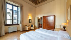 Une chambre d’hôtel avec un grand lit, une chaise, un bureau et une armoire. Il y a une fenêtre avec des rideaux et un radiateur au mur.