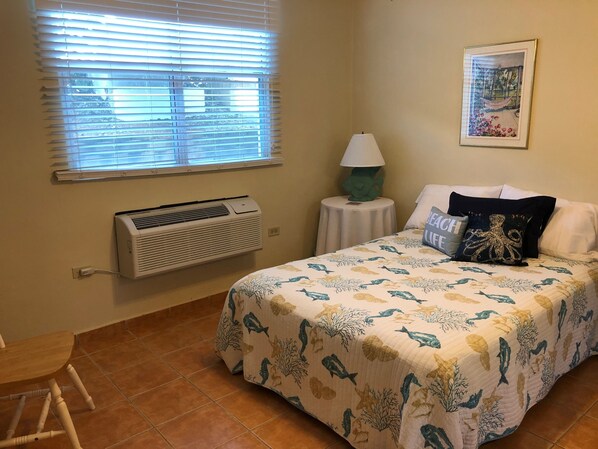 Un dormitorio con cama, ventana con persianas, aire acondicionado, una mesita con lámpara, un cuadro en la pared y una silla.