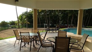 A covered outdoor seating area with tables and chairs, overlooking a swimming pool and palm trees. - Beachfront family friendly garden condo, full A/C, WiFi, sleeps 6, Smart TV's (Cabo Rojo)