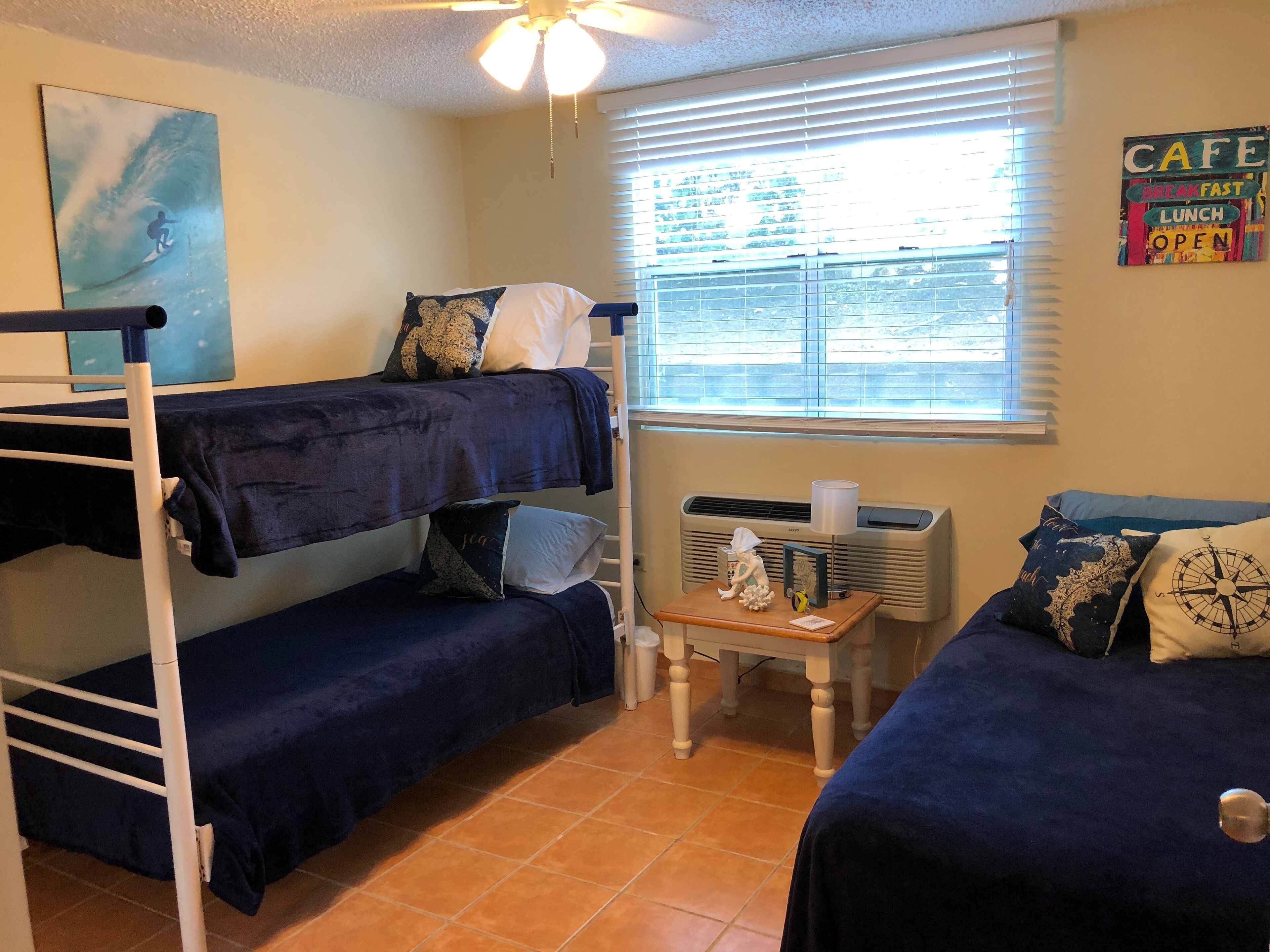 A dormitory room with bunk beds, a heater, a small table with a lamp, and a window with blinds.