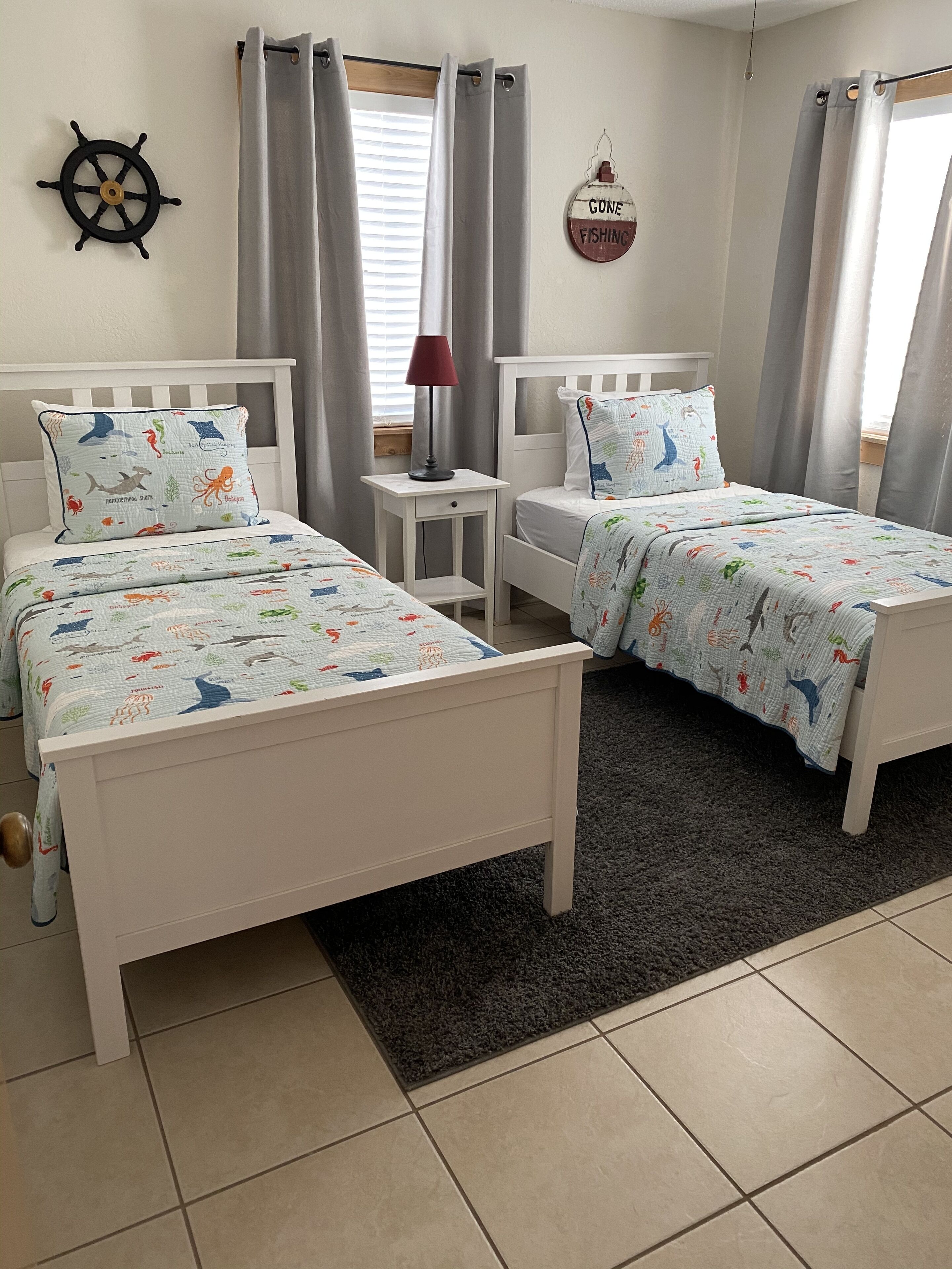 Une chambre avec deux lits, une petite table, une lampe, un décor représentant un gouvernail de bateau et une pancarte indiquant « Pêche au cône ».
