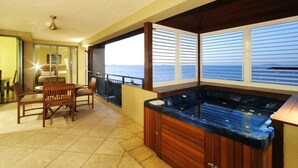 Un balcón con mesa y sillas de madera, jacuzzi y amplias ventanas con vista al mar.