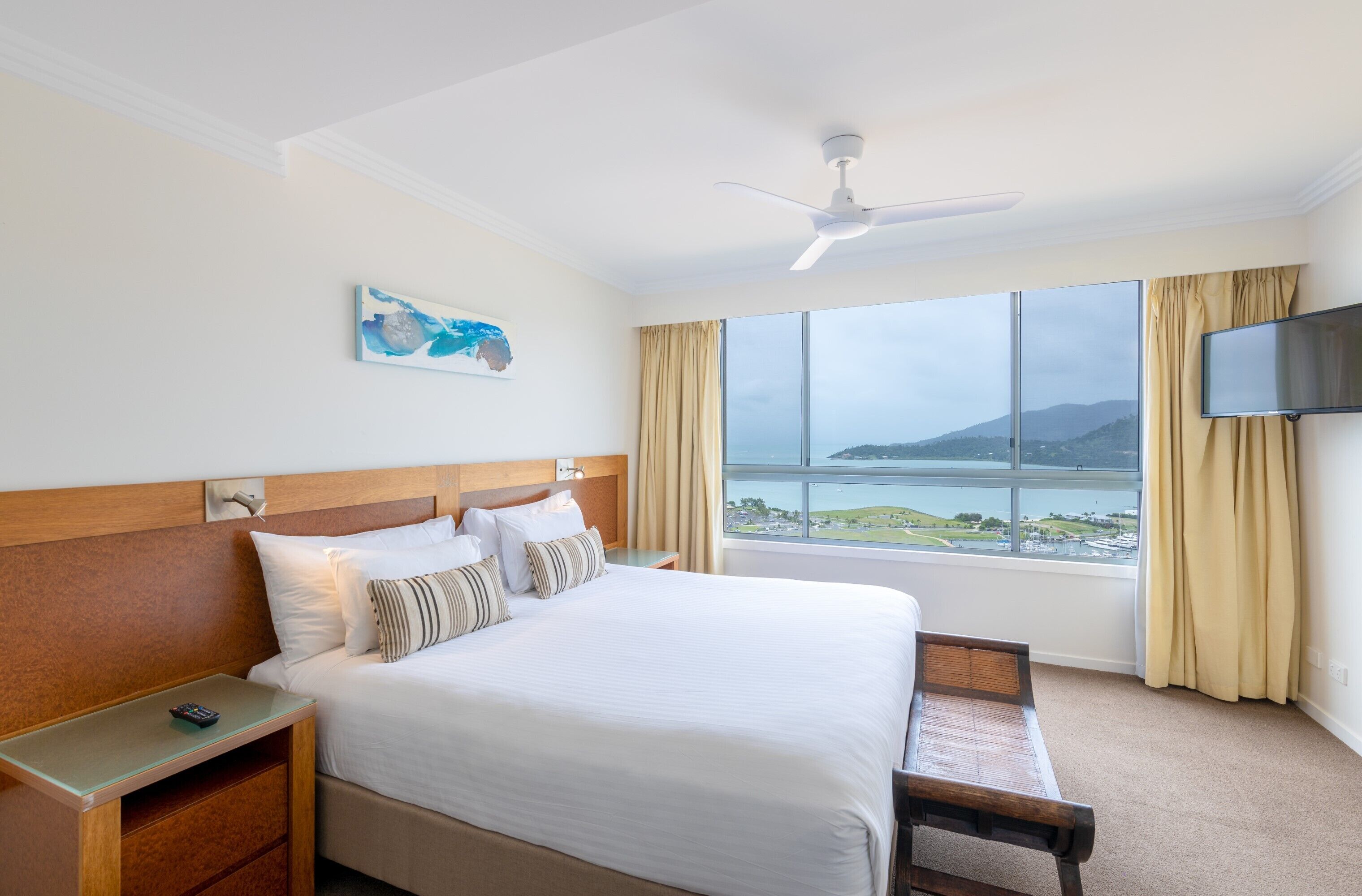 A hotel room with a large bed, a wooden headboard, a bedside table with a remote control, a TV mounted on the wall, and a window with a view of a body of water and hills.