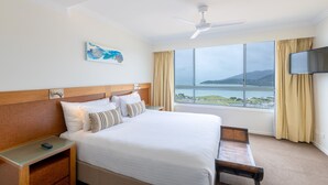 Habitación de hotel con una cama grande, cabecera de madera, mesita de noche con un control remoto, televisión montada en la pared y ventana con vista a un cuerpo de agua y colinas.