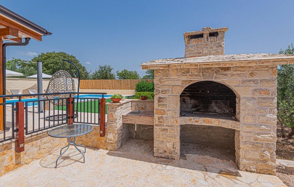 An outdoor patio with a stone-built barbecue area, a glass table, and a pool in the background. - Awesome home in Sibenik (Sibenik)