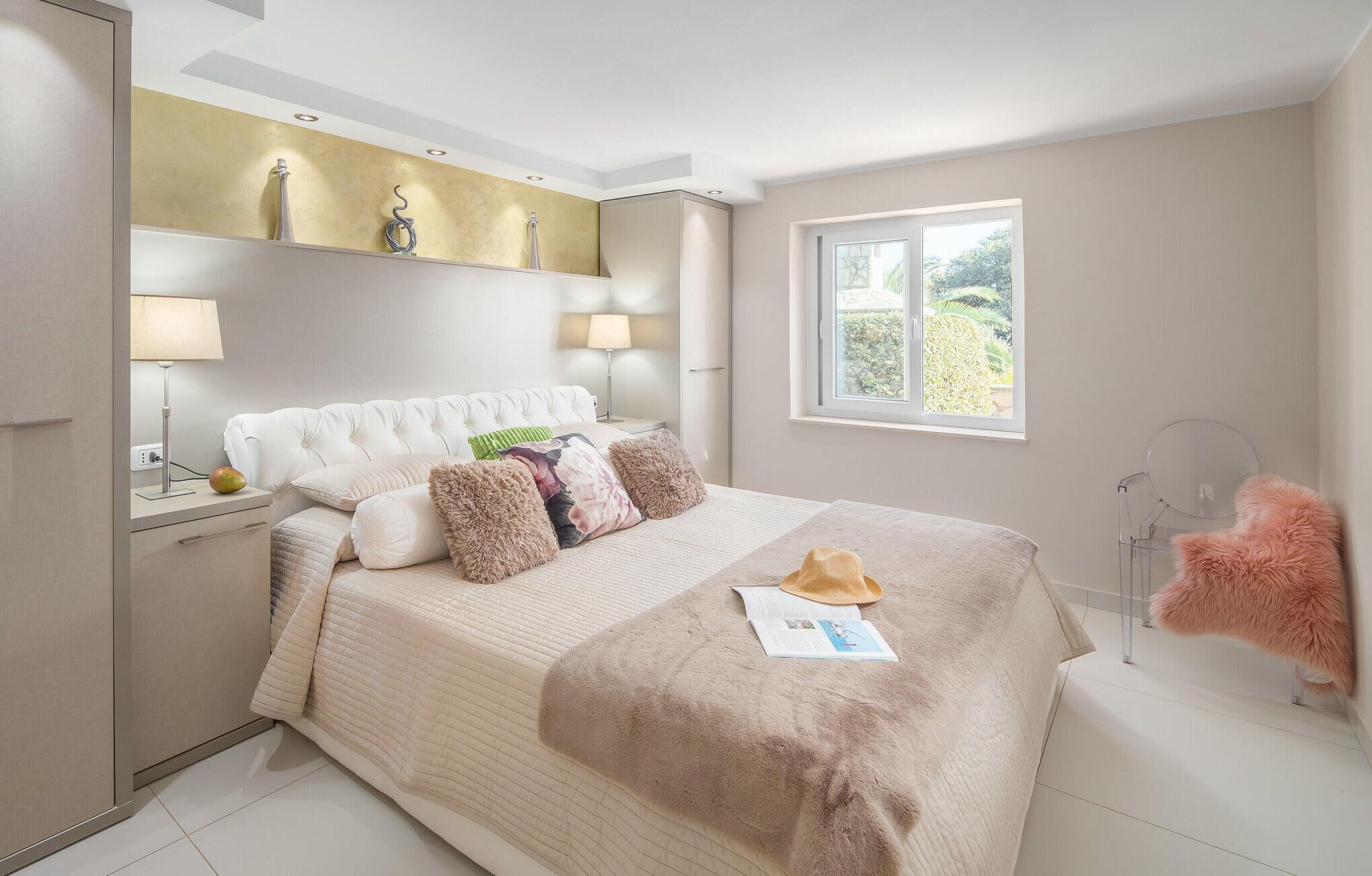 A bedroom with a bed, bedside lamps, a window with a view, and a decorative shelf above the bed.