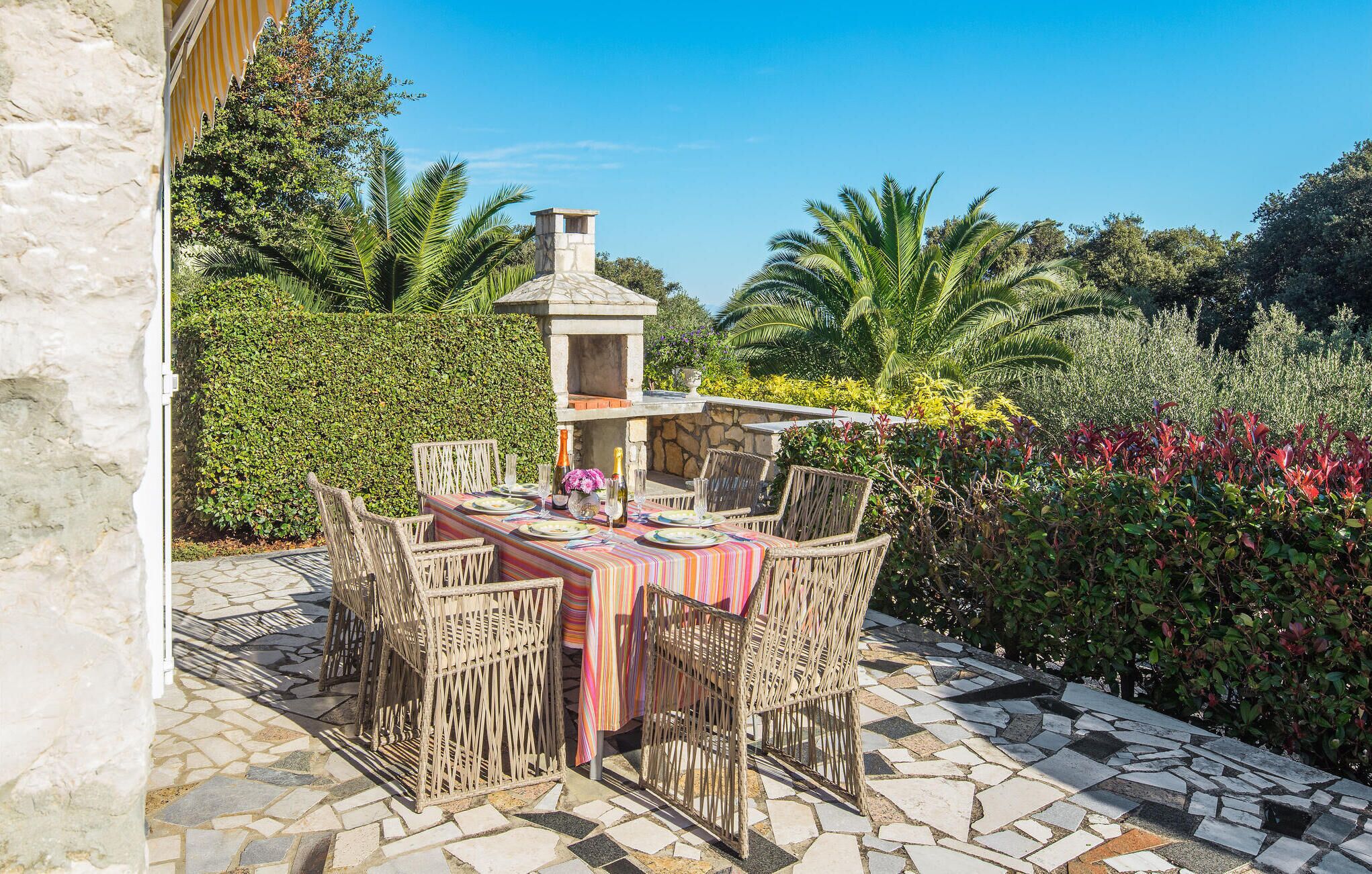 Un espace de restauration en plein air doté d’un patio carrelé en pierre, de chaises en osier et d’une table dressée avec des assiettes et des verres.