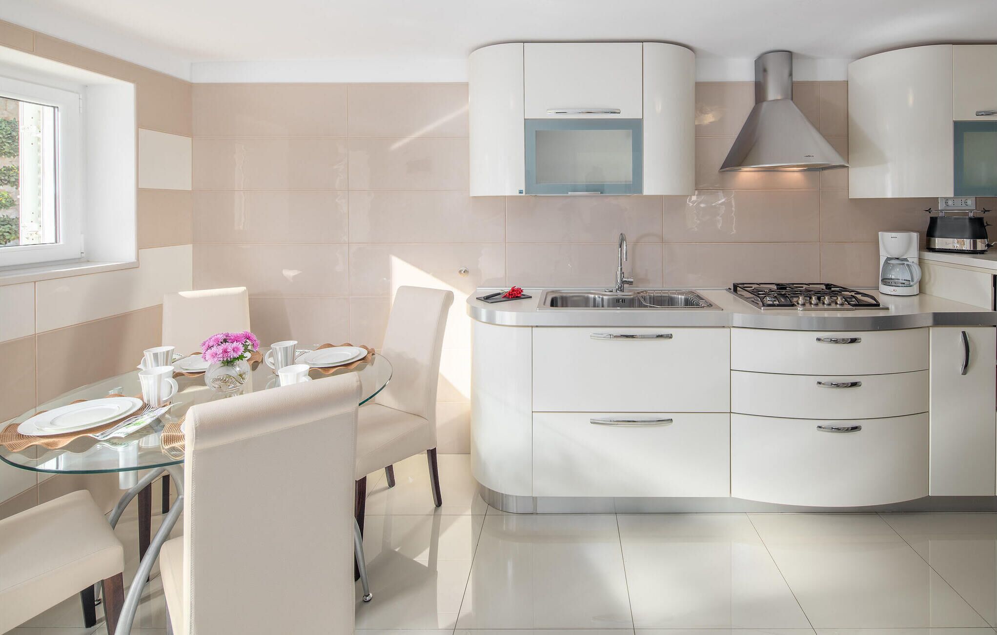 Una cocina moderna con una mesa de comedor de cristal, armarios blancos y un ventanal que proporciona luz natural.