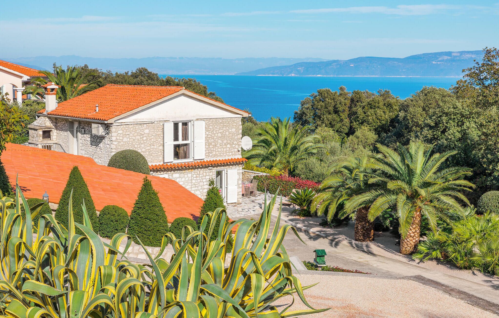 Une maison en pierre, surmontée d’un toit de tuiles rouges, entourée d’une végétation luxuriante et de palmiers, et offrant une vue imprenable sur la côte.