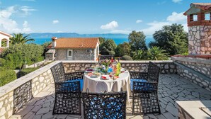 A terrace with a table set for dining, overlooking a coastal view with a stone building and a clear sky. - Lovely home in Ravni with WiFi (Ravni)