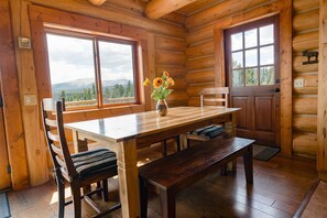 L’intérieur d’un chalet en bois comprend une table à manger, des chaises, un vase avec des fleurs et de grandes fenêtres offrant une vue sur la montagne.