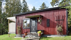 Punainen puinen rakennus, jossa on musta ovi, ikkuna ja pieni kuisti, johon johtaa portaat. Kuistilla on ruukkukasveja ja seinässä metallinen kyltti.