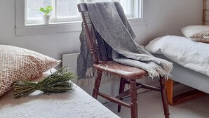 Una habitación con una silla de madera cubierta con una manta gris, una cama con ropa de cama blanca y una ventana con una planta en el alféizar.