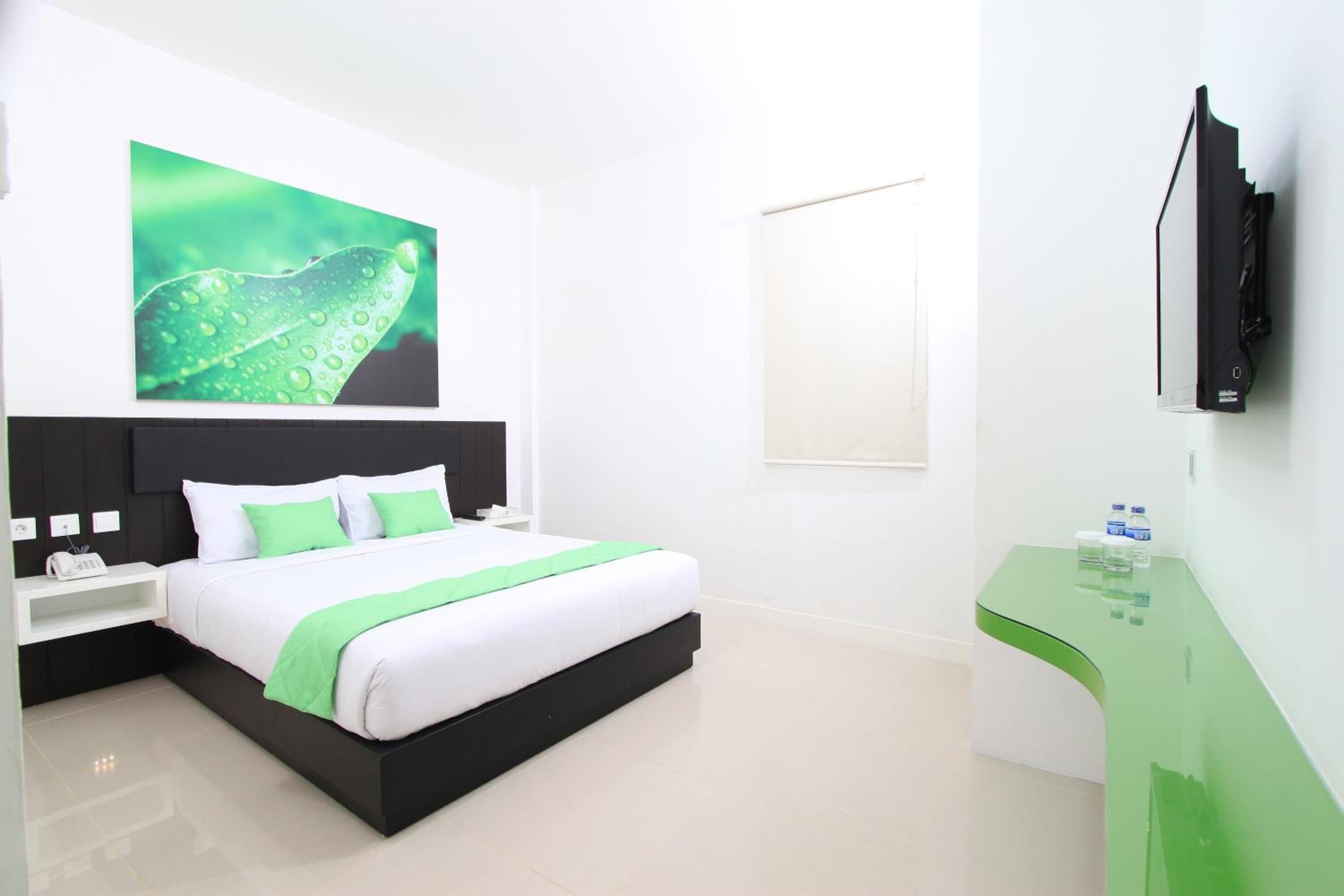 A modern hotel room with a bed, a nightstand, a TV, and a green desk.