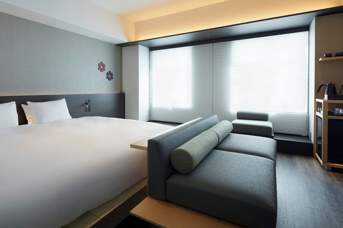 A modern hotel room with a large bed, a sofa, a small table, and a window with blinds.