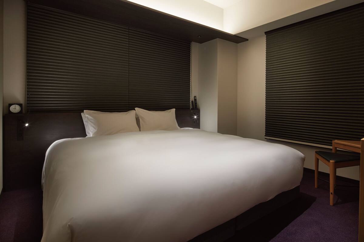 Une chambre d’hôtel moderne avec un grand lit, une tête de lit sombre et des stores occultants.