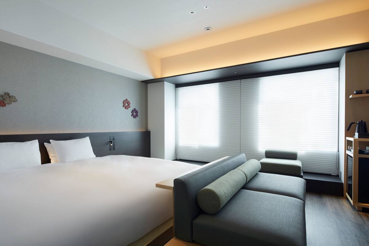Une chambre d’hôtel moderne avec un grand lit, un canapé gris et une fenêtre équipée de stores.