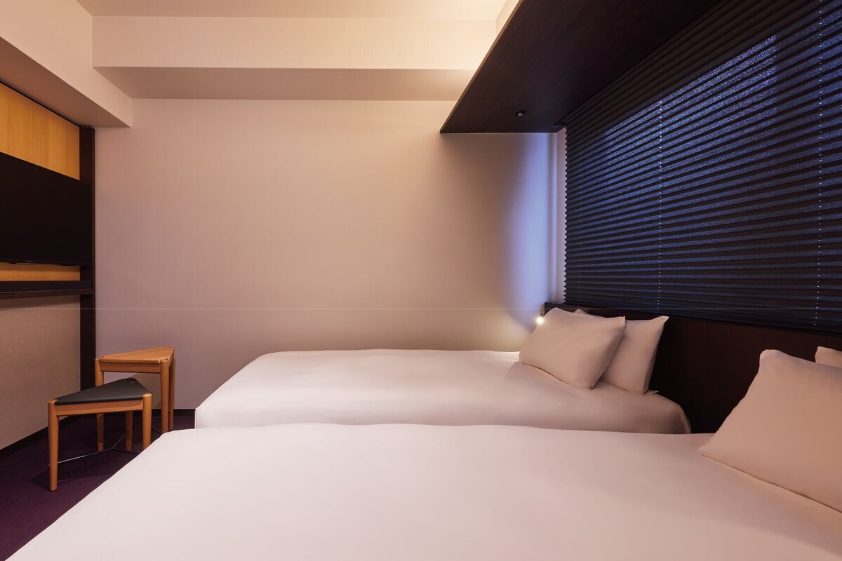 A hotel room with a bed, a TV, a small table, and a window with blinds.