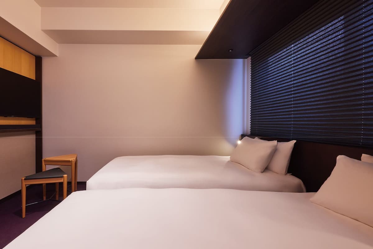 A hotel room with a bed, a TV, a small table, and a window with blinds.