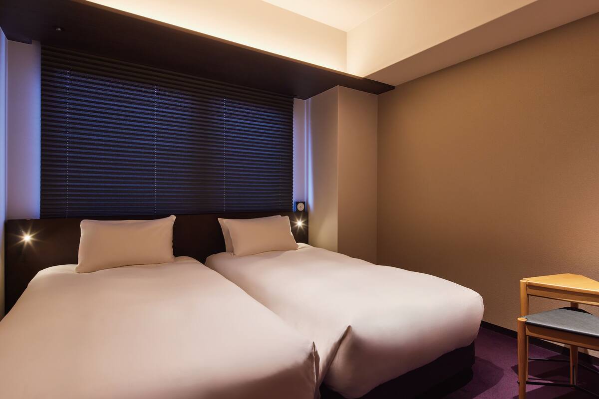 Une chambre d’hôtel avec deux lits, une table de chevet en bois et une fenêtre avec des stores.
